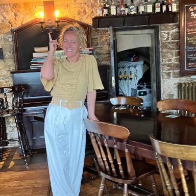 Person standing in a pub with a cheerful expression, wearing casual clothes.